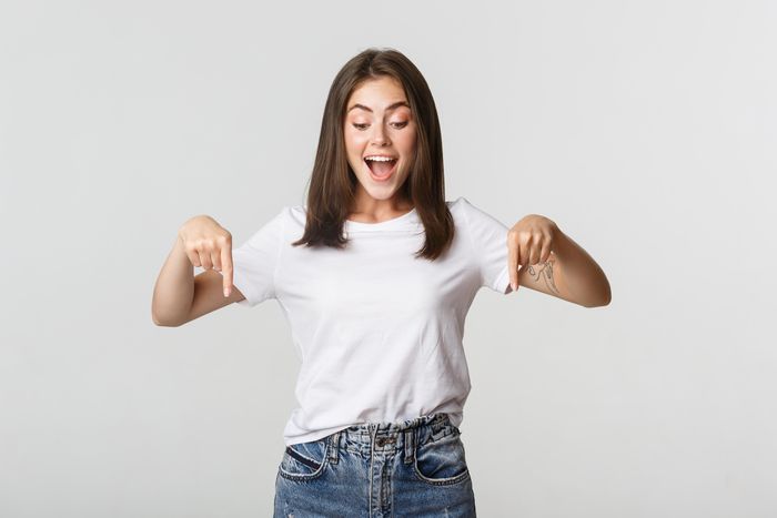 Uma mulher morena, jovem, usando jeans e camiseta branca olha e aponta para baixo com as duas mãos enquanto sorri, ilustra o meu artigo sobre: Cinco maneiras para desenvolver autoestima.