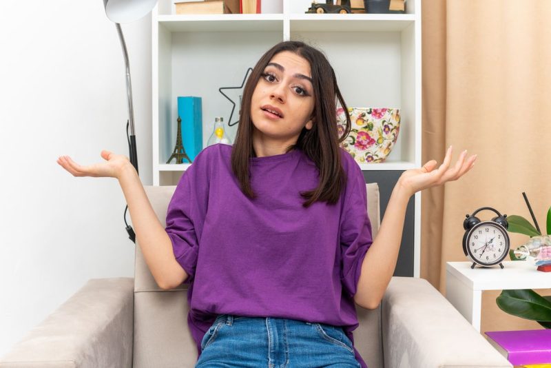 moça, sentada no sofá com as duas mãos um pouco levantadas, simbolizando dúvidas, procurando respostas, usa calça jeans e blusa roxa. Autocuidado.