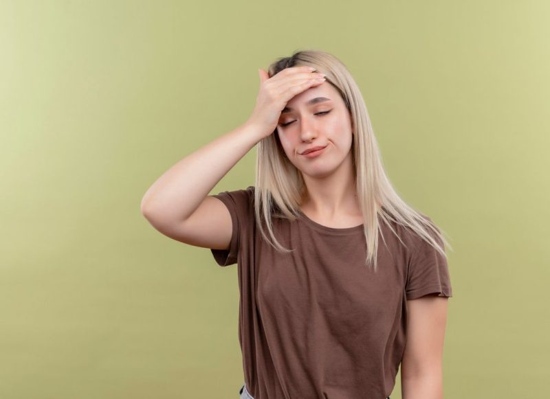 Mulher jovem, loira em pé com as mãos na cabeça. Usa camiseta marrom, está de olhos fechados simbolizando uma desregulação do sistema nervoso.