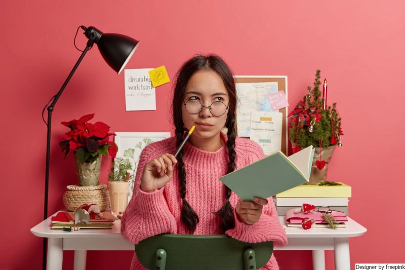 moça, suando óculos e busa de frio rosa. Está com caneta e caderneta nas mãos pensando em suas metas.