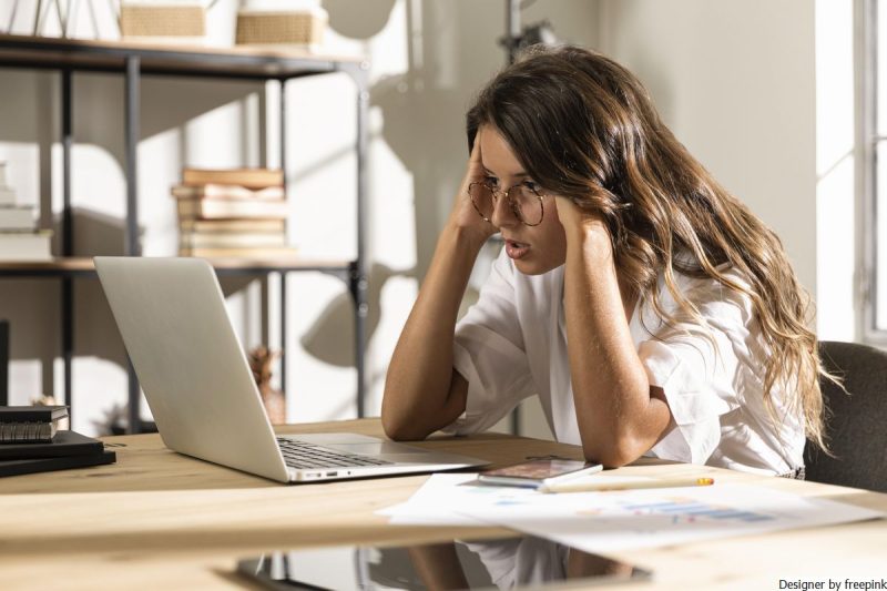 mulher, jovem de óculos com as mãos nas têmporas olha para notbook simbolizando ansiedade de desempenho.