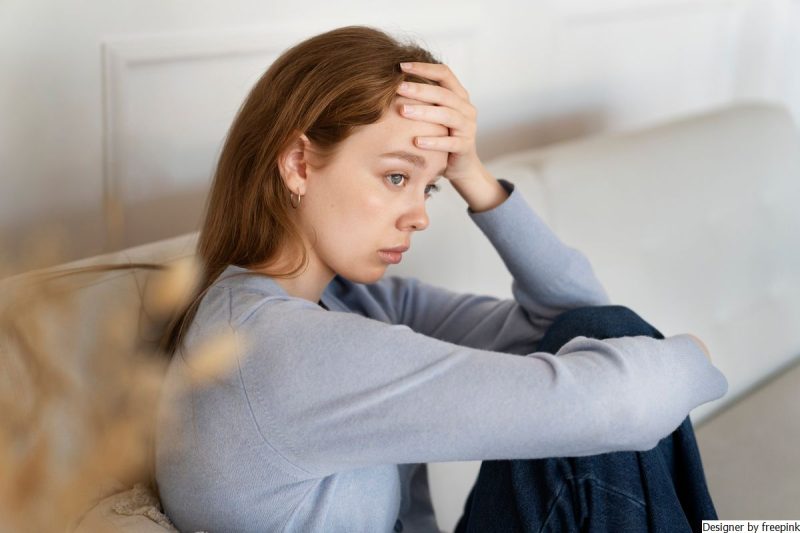 Mulher está sentada no sofá, com mão na cabeça simbolizando tristeza.