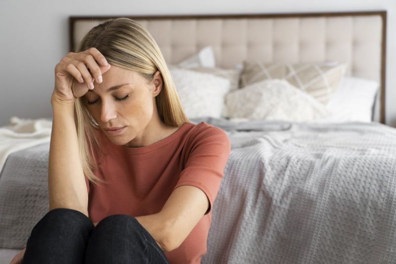 Mulher, jovem, loira está sentada no chão em frente a cama. cabeça baixa uma das mãos na testa, olhos fechados simbolizando esgotamento emocional.
