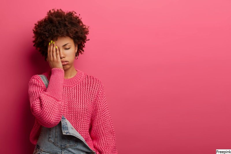 mulher, jovem, usa macacão jeans e blusa de frio de tricô rosa. está com uma das mãos sobre um dos olhos, representando dor emocional.